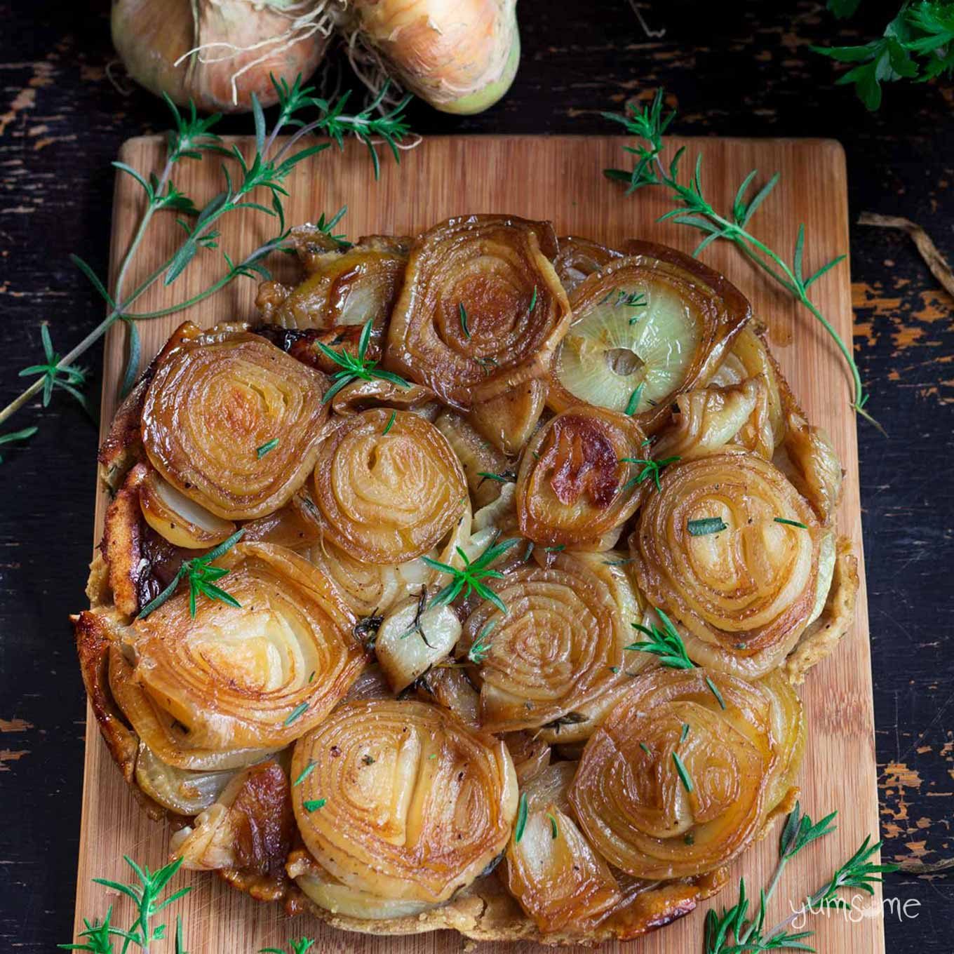 Overhead view of vegan caramelized onion tart on a black table, with some onions and herbs.