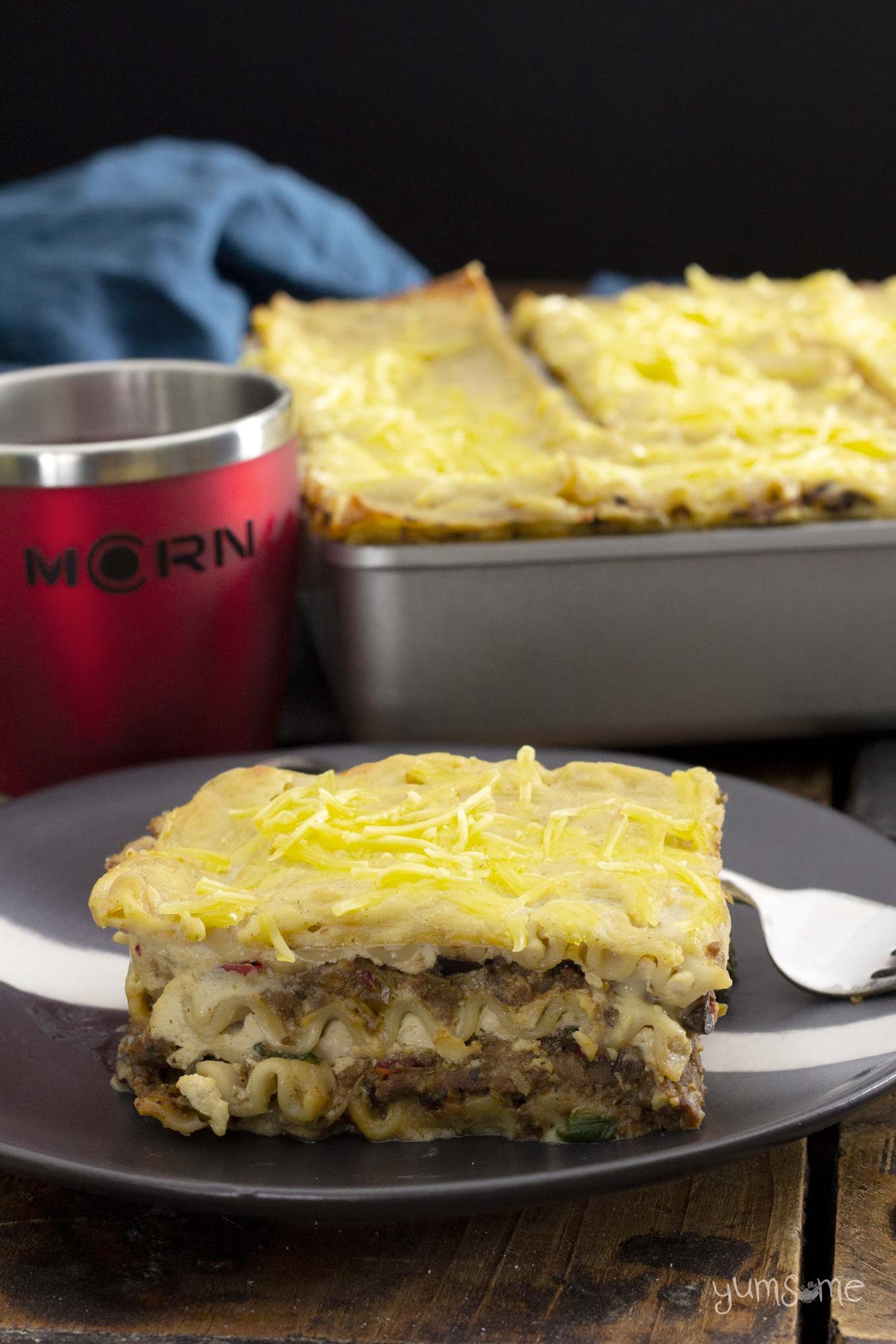 A black plate with a slice of Mariner Valley lasagna, with a tray of lasagna in the background, next to a red MCRN cup.
