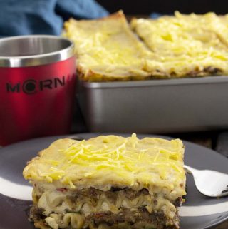 A black plate with a slice of Mariner Valley lasagna, with a tray of lasagna in the background, next to a red MCRN cup.