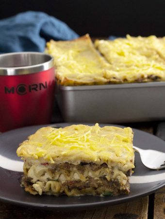 A black plate with a slice of Mariner Valley lasagna, with a tray of lasagna in the background, next to a red MCRN cup.