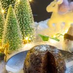 A vegan Christmas pudding, on a black plate, with a slice cut from it.
