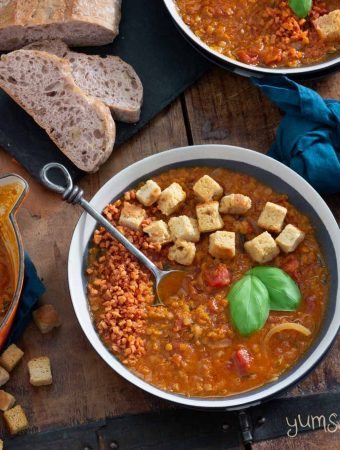 Overhead shot of several bowls of crockpot tomato lentil soup on a wooden table, with croutons scattered around, plus a cut loaf of bread on a black slate.