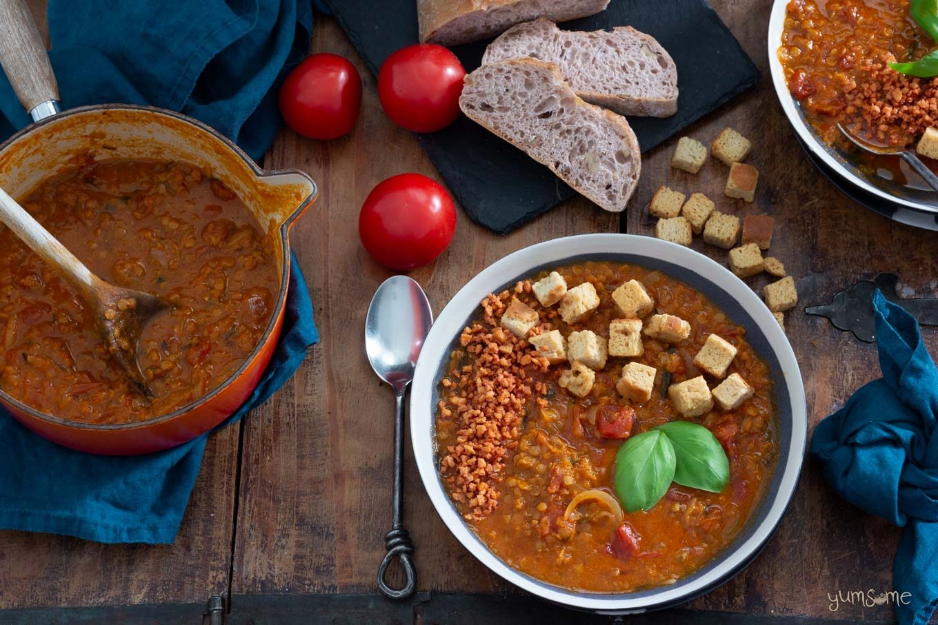 A saucepan of Crockpot Tomato Lentil Soup, plus a black bowl of soup on a wooden table, surrounded by croutons, tomatoes, bread, and blue napkins.