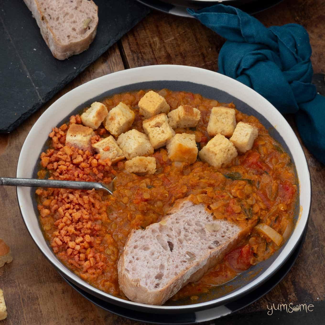 A bowl of Crockpot Tomato Lentil Soup with vegan bacon bits, croutons, and a slice of bread.