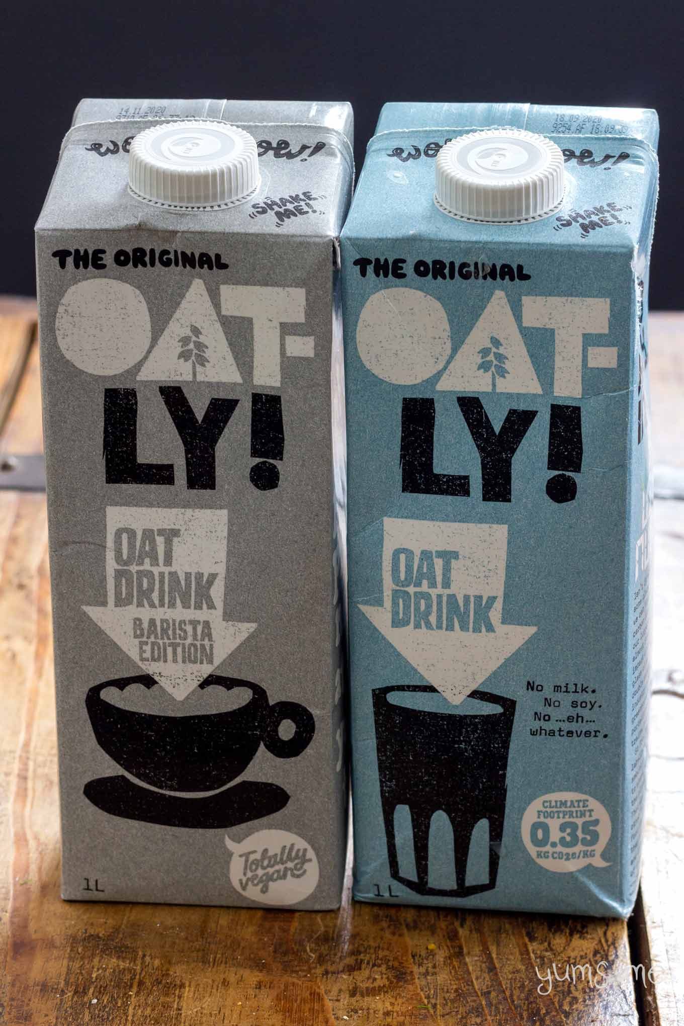 Two cartons of Oatly side by side on a wooden table. Barista (grey), and original (blue).