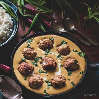 A skillet of malai kofta, plus silver spoons and chillies on a table.