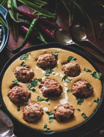 A skillet of malai kofta, plus silver spoons and chillies on a table.