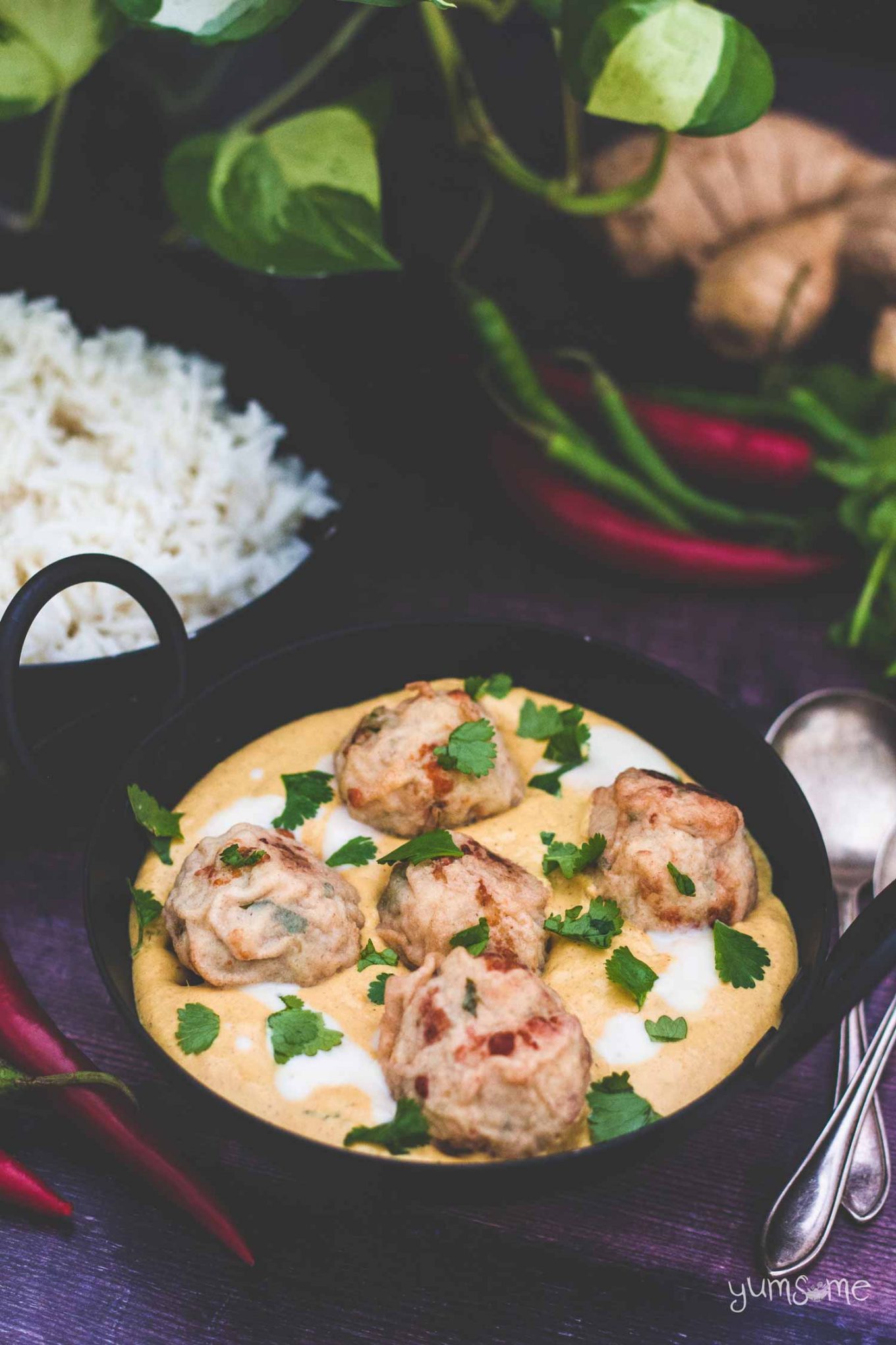 A black bowl of malai kofte with some rice and chillies in the background.